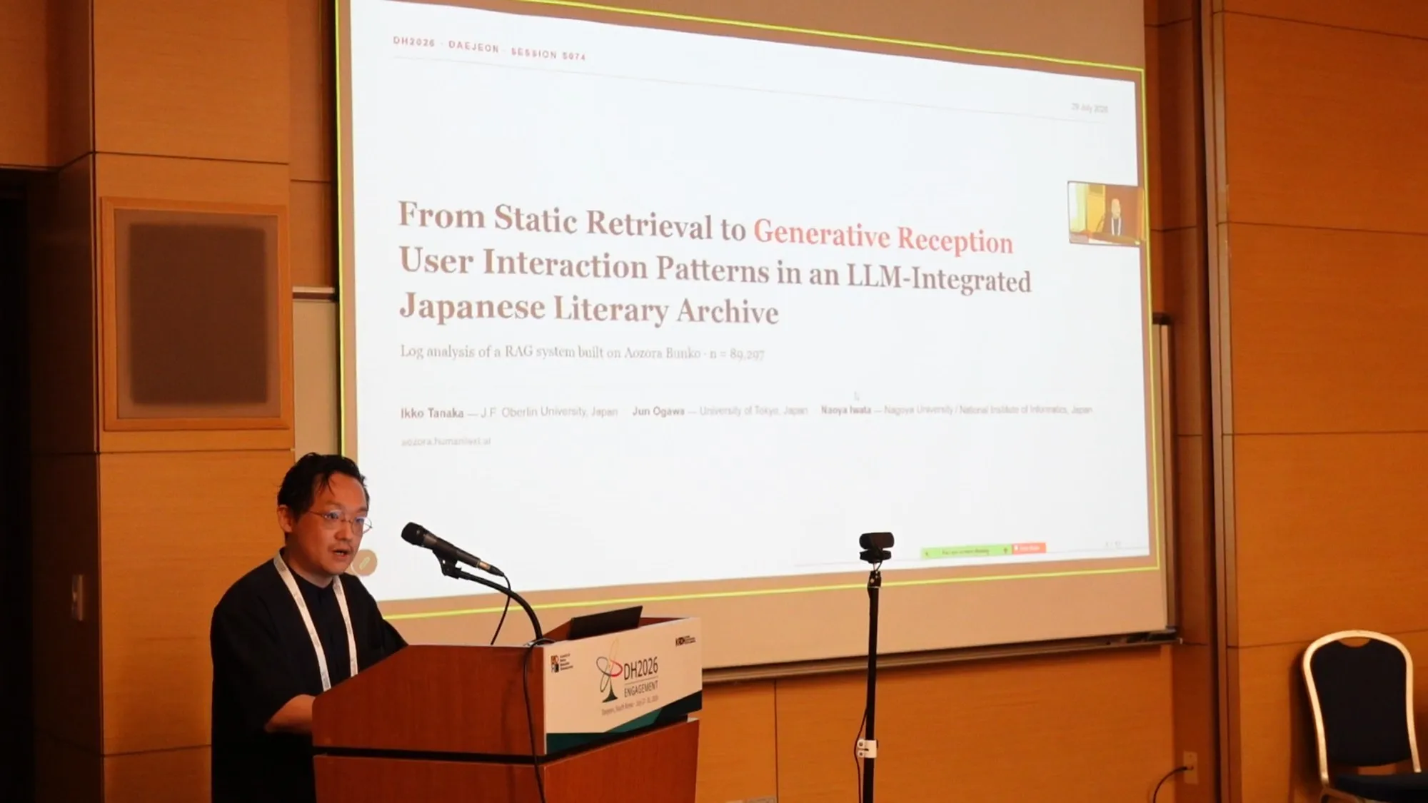 DH2026で発表する田中一孝。スクリーンに「From Static Retrieval to Generative Reception」のタイトルスライド